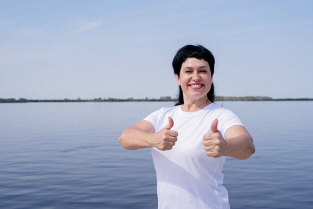 Donna senior attiva e felice in abiti sportivi che mostra i pollici in su che lavora vicino alla riva del fiume