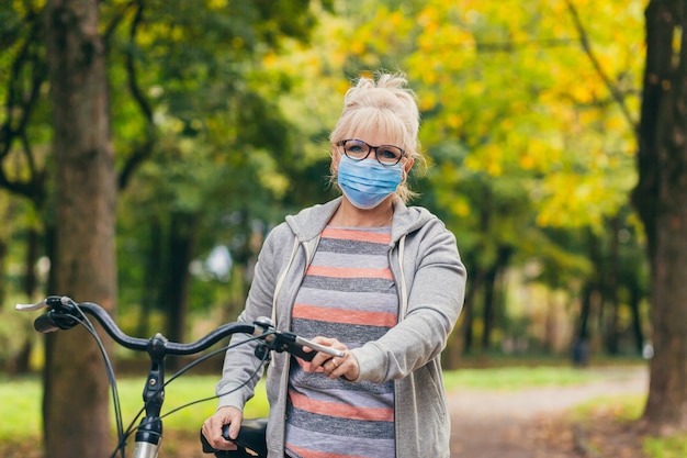 donna senior cammina nel parco con una bicicletta in una maschera medica protettiva