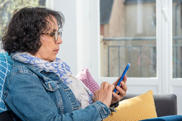 Donna senior castana allegra che per mezzo dello smartphone mentre sedendosi sul sofà