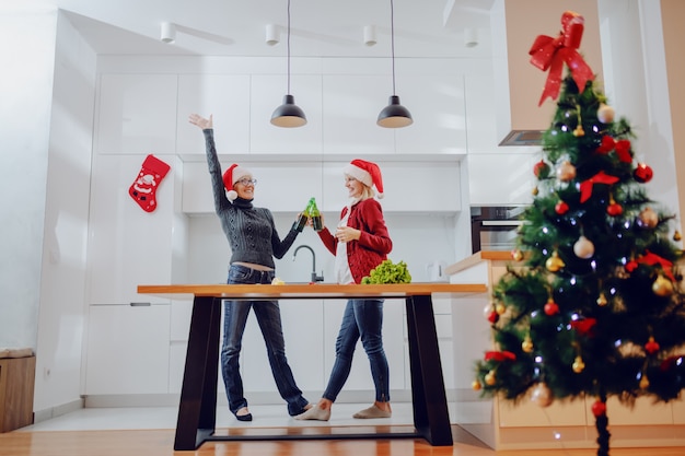 Donna senior caucasica estatica e sua figlia che tostano con la birra in cucina alla notte di Natale. Entrambi hanno cappelli di Babbo Natale in testa. Sul bancone della cucina ci sono verdure.