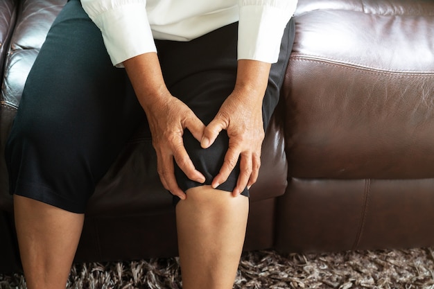Donna senior che soffre dal dolore al ginocchio a casa, concetto di problema sanitario