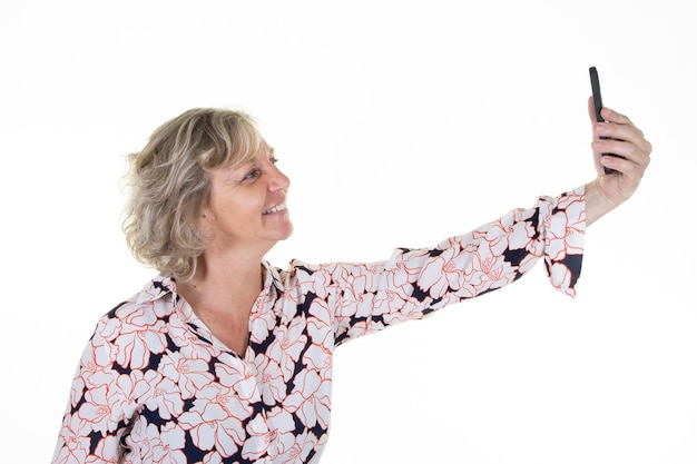 Donna senior felice gioiosa che tiene telefono cellulare e sorridente prendendo selfie felice isolato su sfondo bianco