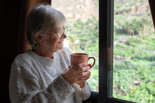 Donna senior pensierosa sorridente che tiene una tazza del tè o del caffè che guarda fuori dalla finestra