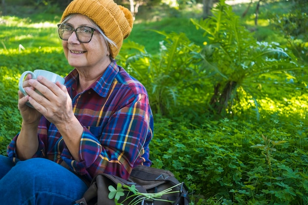 Donna senior rilassata con cappello giallo che tiene una tazza di tè seduta nel prato