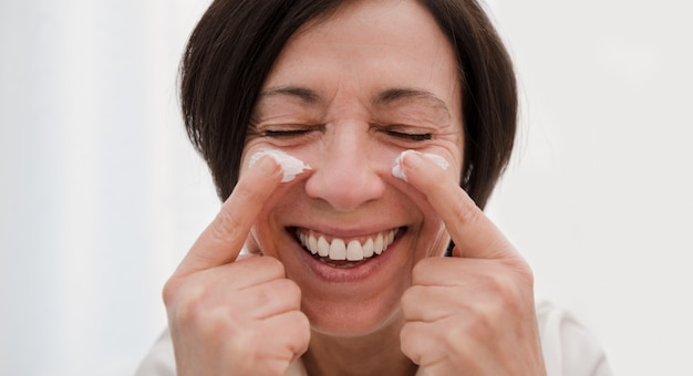 Donna senior sorridente che applica la crema di fronte antinvecchiamento a casa. Donna matura felice che usando idratante di giorno