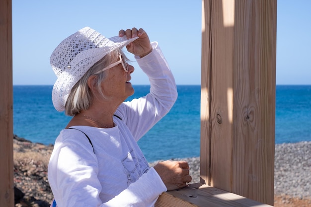 Donna senior sorridente felice in bianco che indossa cappello e occhiali da sole guardando l'orizzonte sul mare