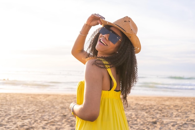 Donna sensuale che indossa occhiali da sole e cappello da sole in posa sorridendo alla telecamera sulla spiaggia