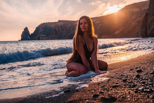 Donna sensuale spensierata felice del fuoco selettivo con i capelli lunghi in costume da bagno nero che posa alla spiaggia del tramonto
