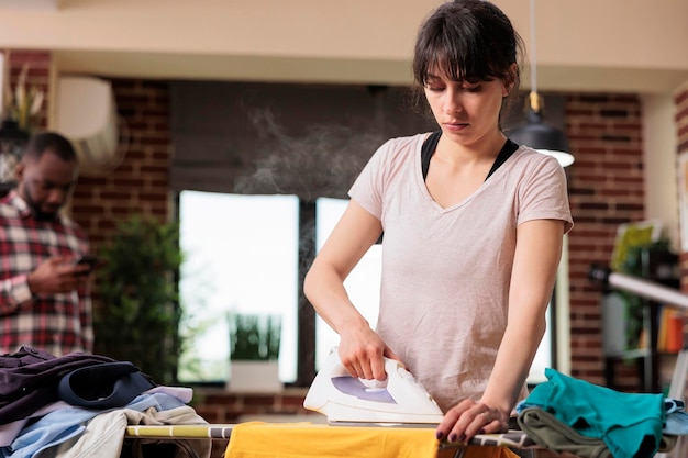 Donna seria che si concentra sulla stiratura dei vestiti a casa mentre il fidanzato controlla i messaggi sul cellulare in background. Moderna coppia multirazziale che trascorre del tempo insieme a casa, faccende domestiche.