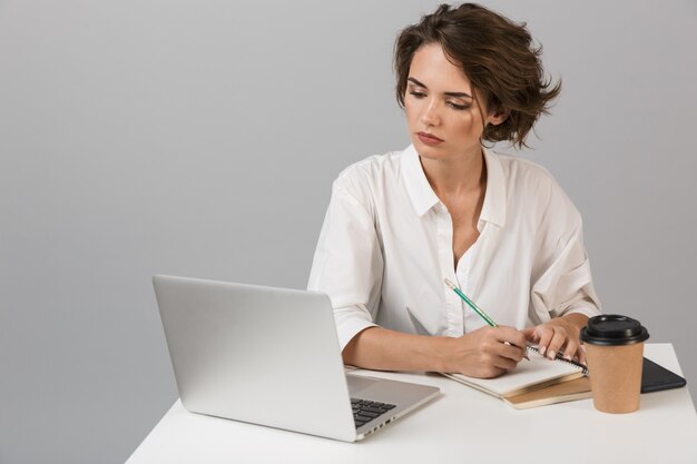 Donna seria di affari che posa isolata sopra la parete grigia che si siede al tavolo utilizzando il computer portatile