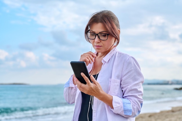 Donna seria triste di mezza età in cuffie con smartphone all'aperto