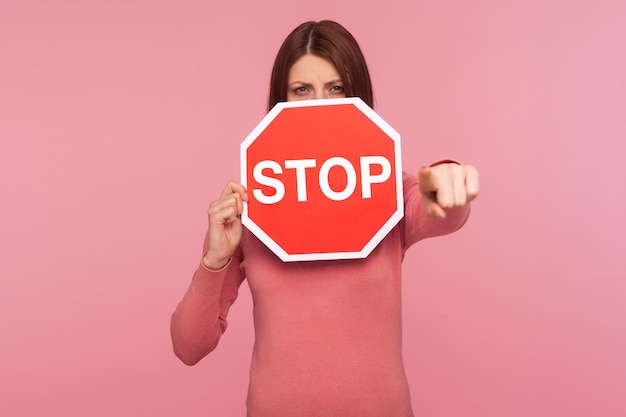 Donna severa arrabbiata in maglione rosa che tiene il segnale di avvertimento di arresto e punta il dito verso di te accedi al divieto proibito Colpo in studio al coperto isolato su sfondo rosa