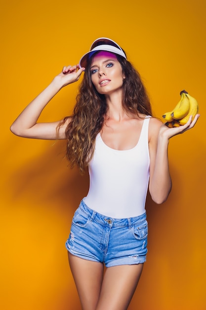 Donna sexy in costume da bagno bianco e pantaloncini di jeans blu, visiera alla moda che tiene le banane e posa isolate sopra il giallo