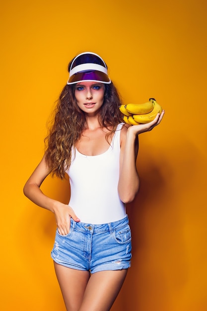 Donna sexy in costume da bagno bianco e pantaloncini di jeans blu, visiera alla moda che tiene le banane e posa isolate sopra il giallo