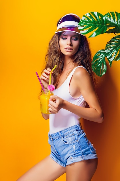 Donna sexy in costume da bagno bianco e pantaloncini di jeans blu, visiera alla moda che tiene un bicchiere di bevanda fresca mentre si sta in piedi sul giallo, arancione caldo