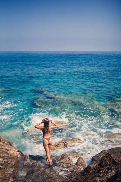 Donna sexy in costume da bagno completo con capelli lunghi cammina su grandi rocce su una spiaggia rocciosa durante una tempesta in mare Vista posteriore Costume da bagno donna isola tropici paesaggio passeggiata esotica