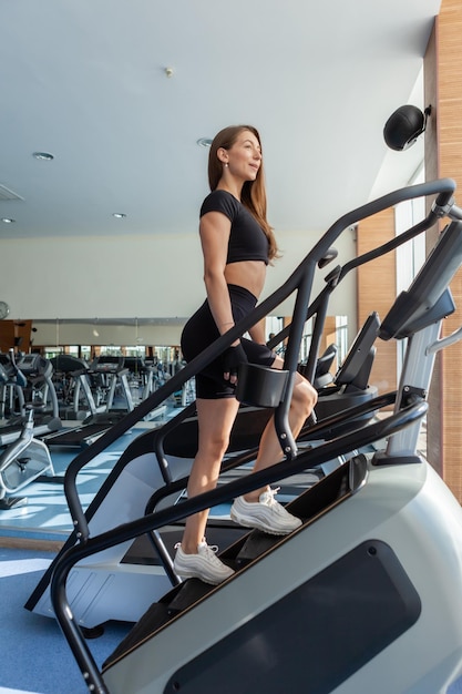 Donna sexy in forma in abiti sportivi che fa esercizio cardio su una macchina per esercizi a gradini in una palestra moderna con grandi finestre