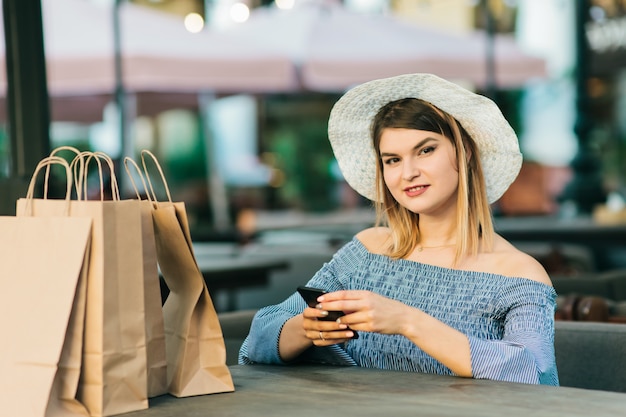 donna shopaholic in elegante abito e cappello si siede al tavolo nella caffetteria all'aperto e utilizza uno smartphone