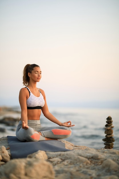 Donna simile allo Zen che pratica Yoga con gli occhi chiusi al mattino su una roccia in riva al mare.