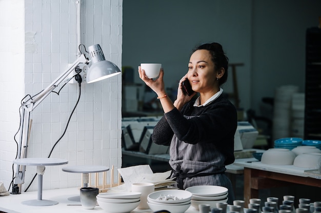 Donna sociale esaminando una tazza sotto una lampada su un tavolo con stoviglie e strumenti in bianco di recente produzione