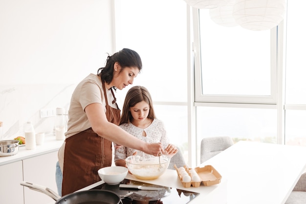 Donna soddisfatta soddisfatta e piccola figlia che cuociono pasticceria in cucina a casa e che impastano la pasta con il cucchiaio