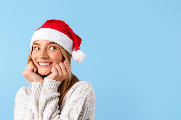 Donna sognante con cappello da Babbo Natale che guarda lo spazio libero e sorride su sfondo blu studio