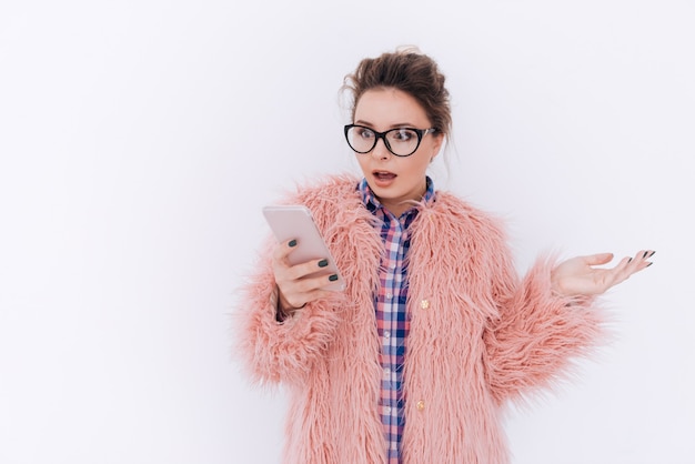 Donna sorpresa con gli occhiali e la pelliccia rosa che tiene il telefono e lo guarda. Muro grigio isolato