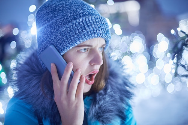 Donna sorpresa vestita in abiti invernali, apre la bocca con stupore, parla con il suo ragazzo tramite smartphone