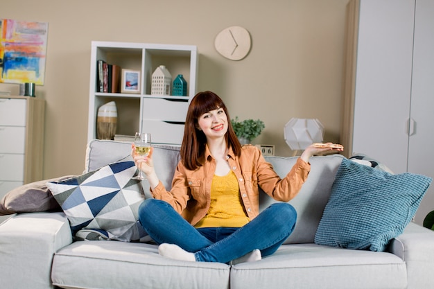 Donna sorridente abbastanza giovane in jeans e camicia gialla, bevendo vino e mangiando pizza, mentre sedendosi sul sofà grigio a casa