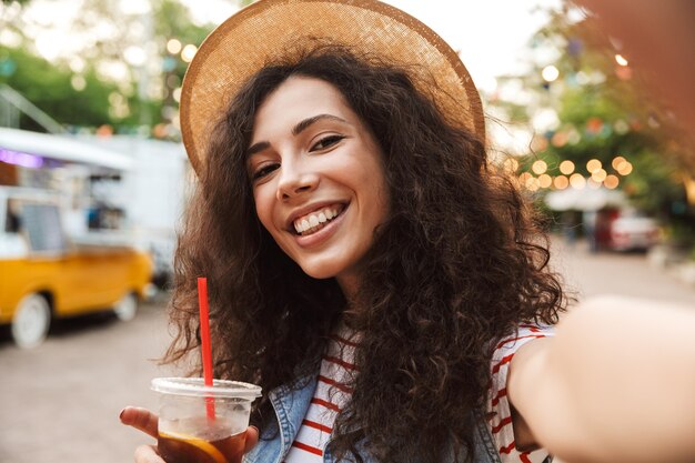 donna sorridente affabile con capelli castani ricci che indossa un cappello di paglia estivo sorridente e bere tè freddo da un bicchiere di plastica all'aperto, mentre scatta foto selfie