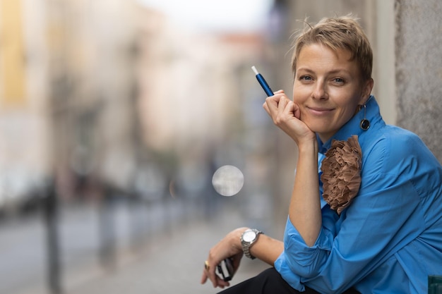 Donna sorridente alla moda adulta che si siede sulla via europea e che tiene un looki della sigaretta elettronica