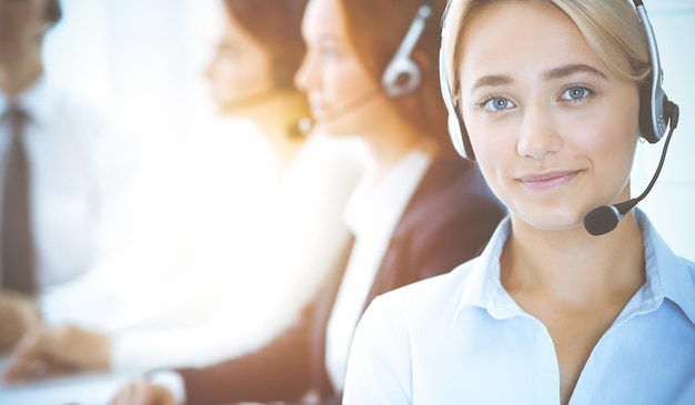 Donna sorridente allegra di affari con le cuffie che consultano i clienti. Gruppo di diversi operatori telefonici al lavoro in ufficio soleggiato. Concetto di call center e uomini d'affari.