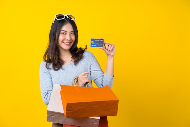 Donna sorridente asiatica felice che presenta la carta di credito e che porta una borsa coloful di compera per la presentazione dell'acquisto online sulla parete gialla isolata