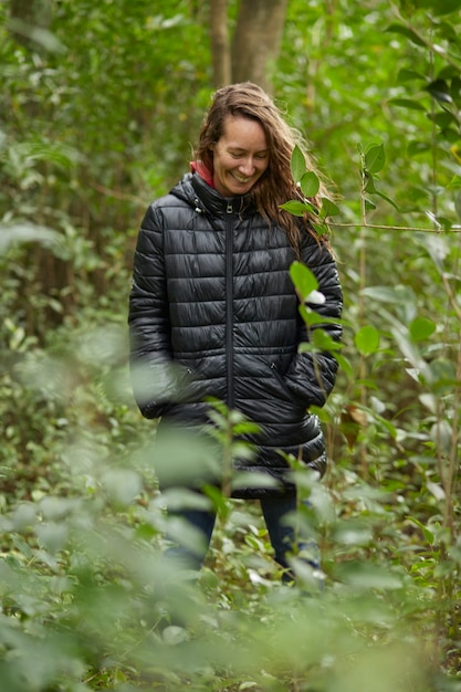 Donna sorridente bionda di mezza età che cammina nella boscaglia in inverno, tra le foglie degli alberi. Indossa una giacca calda.