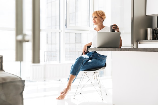 Donna sorridente casualy vestito utilizzando il computer portatile mentre era seduto su una cucina, indossando gli auricolari