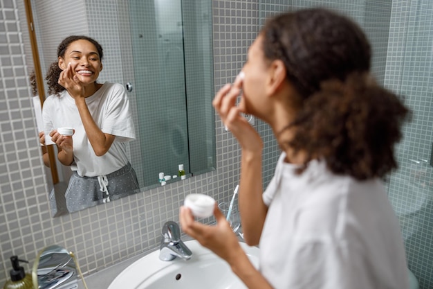 Donna sorridente che applica crema antirughe in piedi dietro lo specchio nella procedura di bellezza del bagno di casa
