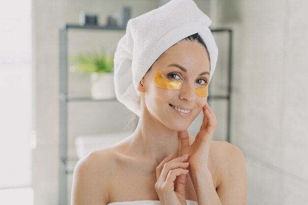 Donna sorridente che indossa un asciugamano con cerotti idrogel sulla pelle sotto gli occhi in bagno Routine di cura della pelle