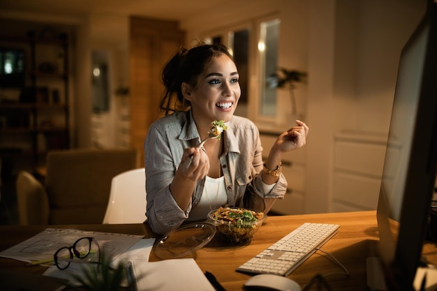 Donna sorridente che legge un'e-mail sul PC desktop e mangia insalata la sera a casa