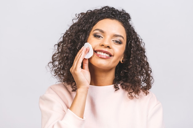 Donna sorridente che rimuove il trucco. La foto della donna afroamericana tiene i tamponi di cotone vicino al fronte isolato su priorità bassa bianca. La cura della pelle e il concetto di bellezza.