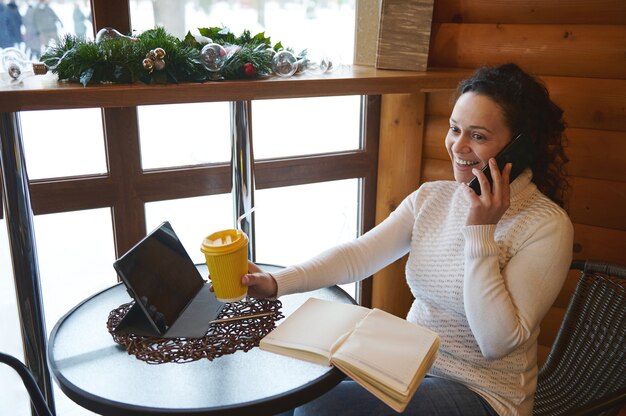 Donna sorridente che tiene il bicchiere di carta del caffè e parla dal telefono cellulare mentre era seduto al bar Affari al caffè.