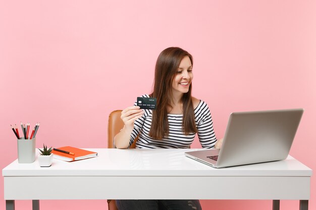 Donna sorridente che tiene la carta di credito mentre lavora al progetto mentre è seduta in ufficio con il laptop