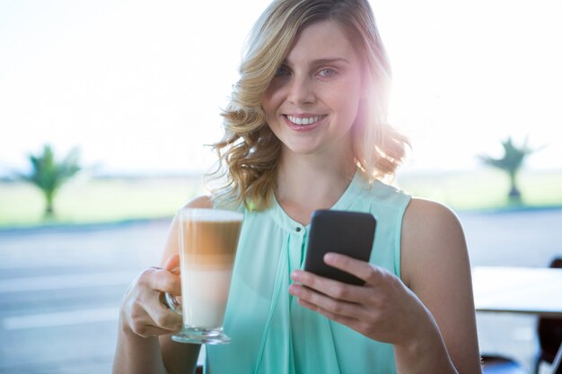 Donna sorridente che tiene un bicchiere di caffè e che per mezzo del suo telefono cellulare