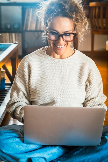 Donna sorridente che usa un portatile che controlla le notizie via e-mail online che siede sul pavimento che cerca amici su social network o che lavora al computer che scrive un blog o che guarda un webinar che studia a casa