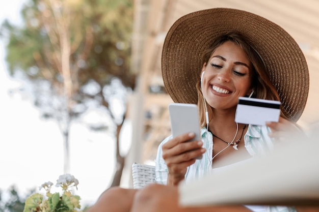 Donna sorridente che utilizza il telefono cellulare e la carta di credito per lo shopping online mentre ci si rilassa in un bar