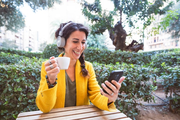 Donna sorridente con cuffie wireless con un caffè in mano e guardando il telefono