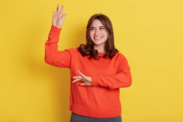 Donna sorridente con felice espressione facciale che guarda sognante da parte, alzando le mani e ballando, celebrando il suo successo, in posa isolata sopra il muro giallo.