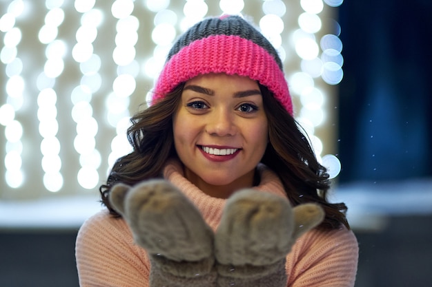 Donna sorridente con ghirlande e luci natalizie in fiera di Natale o Capodanno. Signora che indossa maglione e guanti lavorati a maglia classici alla moda