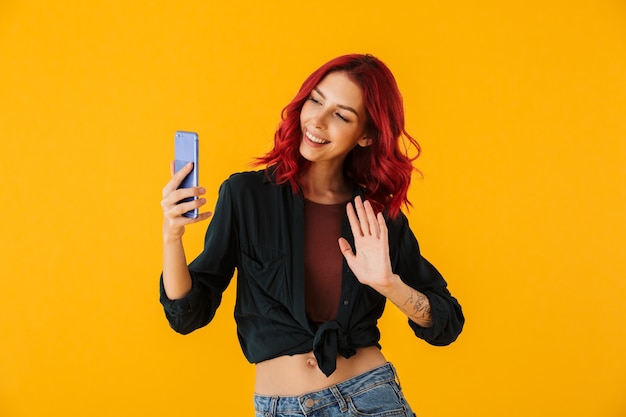 donna sorridente con i capelli rossi ricci agitando la mano e prendendo selfie foto sul cellulare isolato