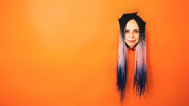 Donna sorridente con i dreadlocks che guarda l'obbiettivo Giovane femmina allegra con capelli multicolori che guarda l'obbiettivo nel buco su sfondo arancione in studio