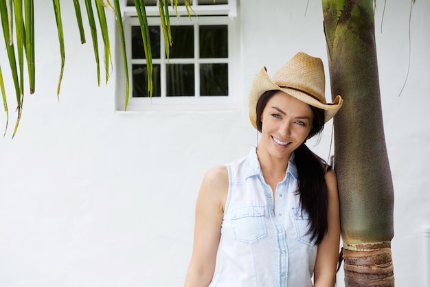 Donna sorridente con il cappello da cowboy che si appoggia contro l&#39;albero